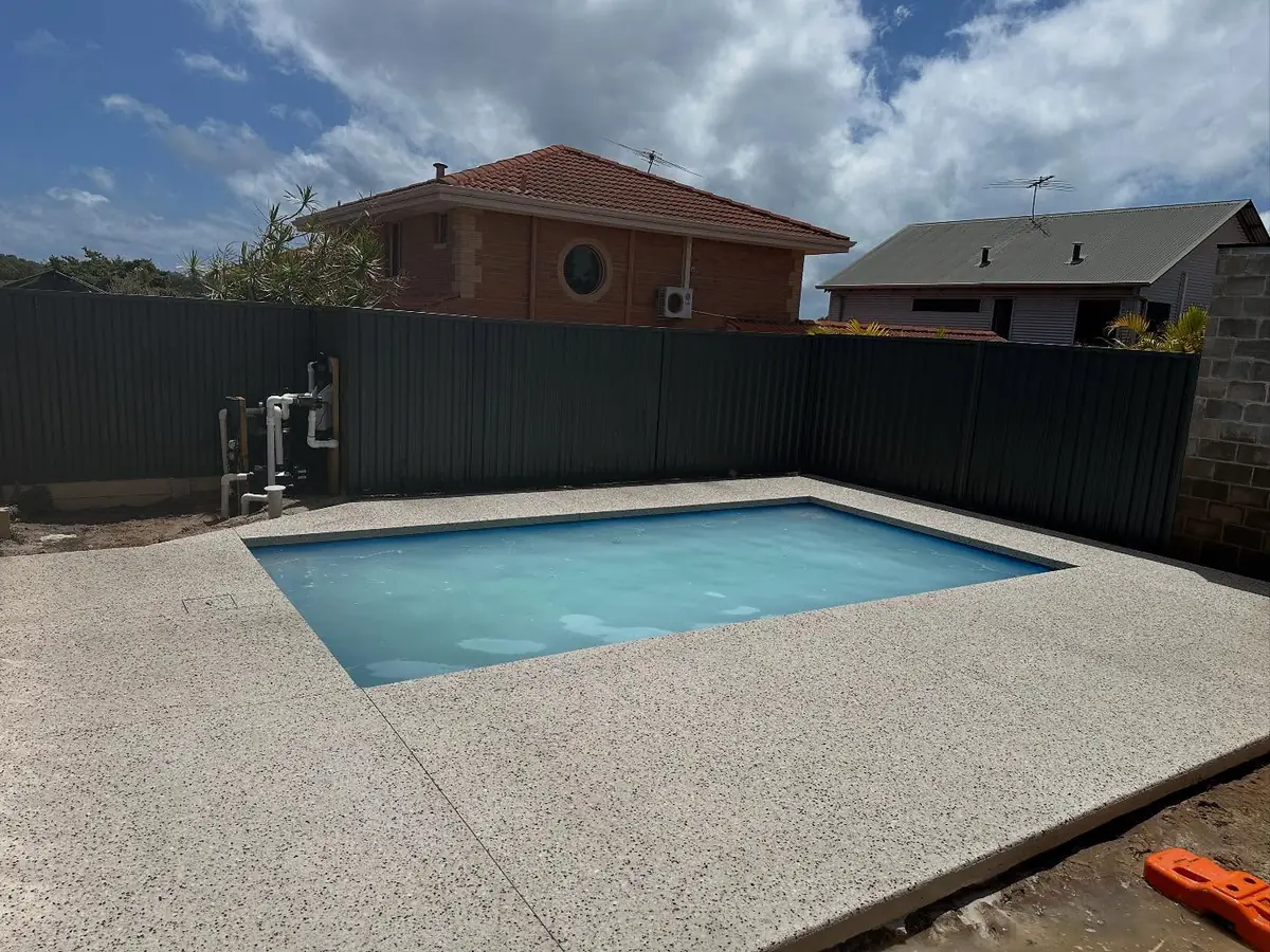 Light exposed aggregate surround around a rectangular pool with the fence and neighbouring houses behind