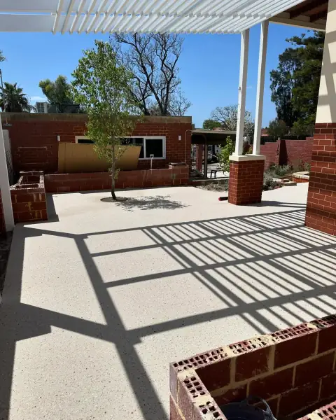 Exposed aggregate courtyard floor under a white pergola with brick planters, a young tree in the middle