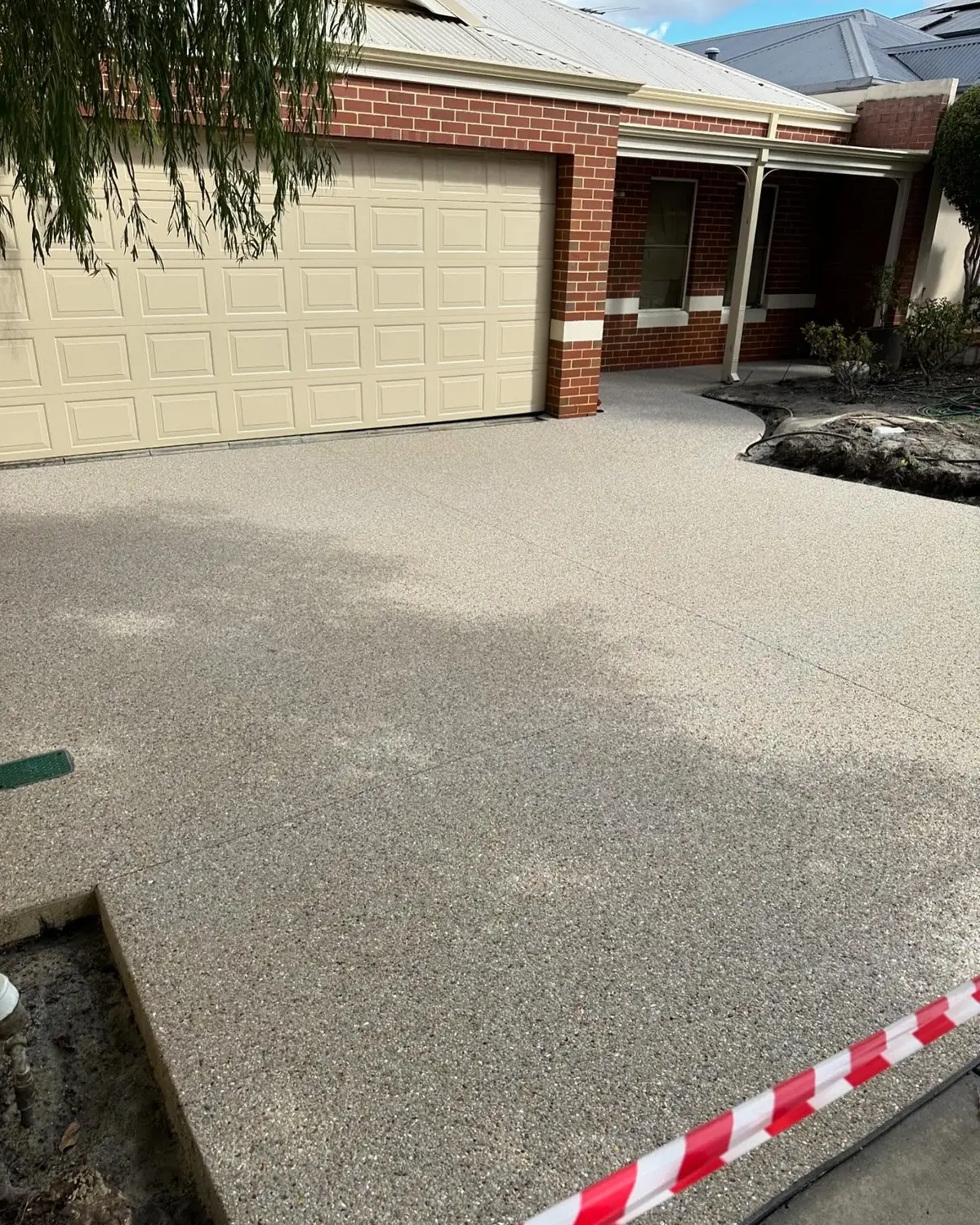 Exposed aggregate driveway in front of a brick home with a cream garage door, taped off while it cures